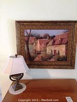 Photo showing wooden framed painting of village houses above the table with lamp.