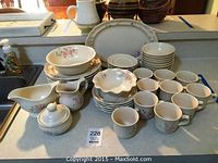 Photo showing full collection arranged on a counter including mugs, plates, bowls, cream and sugar, and covered container, floral design visible.