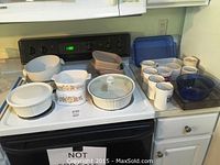 Wide view of all bakeware, casserole dishes, and mugs arranged on kitchen counters and stove.