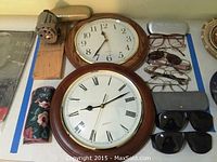 Photo showing two quartz wall clocks with wooden frames, six pairs of eyeglasses with two cases, two pairs of dark solar shield sunglasses, and a vintage hand cranked pencil sharpener mounted on wood.