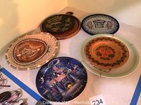 Seven decorative plates on display showing Disneyland plate with castle and fireworks, various themed decorative plates in wood, metal, and ceramic.
