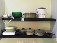Photo of two black shelves holding a variety of kitchen cookware including pots, pans, a green glass bowl, an electric hand mixer, and a white Rival crock pot with blue floral pattern.