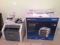 Photo showing the Brother MFC-9130cw printer next to its original box, with some cables and accessories on top of the printer.