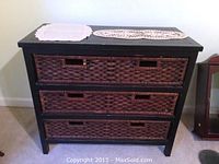 Front view of the black wood chest with three reddish-brown woven wicker drawers and lace doilies on top.