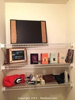 Three wire shelves filled with various items: large leather-bound scrapbook/album with painted tropical scene, a row of books including Bruce Wilkinson titles, a red bean-shaped pillow with UGA logo, assorted small bags, a fanny pack, water bottle, and small containers.