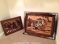 Two framed wooden religious art pieces leaning on floor; one showing Jesus and children outdoors, larger with floral border; other showing three women gathering water inside home