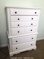 Front view of the white wood chest of drawers showing nine drawers with black knobs, decorative corner grooves, and scalloped base.