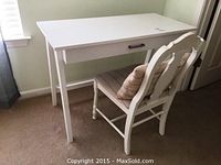 Front view of white wood desk with drawer and silver handle beside white wood chair with cushioned seat.