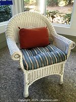 Front angle view of white wicker patio chair with striped blue and beige seat cushion and rust-colored back pillow on a porch.
