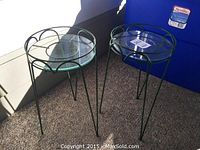 Two round metal patio end tables with clear glass tops and thin angled legs on carpeted floor.