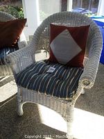 Front view of white wicker chair showing armrest, striped seat cushion, and red and white pillow.