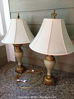 Two matching ornate table lamps with white fabric shades and gold-toned decorative bases on carpeted floor, side by side
