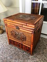 Front left angle view of the wood storage box showing carved palm tree plaque, latch, and woven panel sides.
