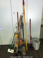 Photo showing collection of brooms, cleaning tools, garden hose and watering can against white cabinet door background.