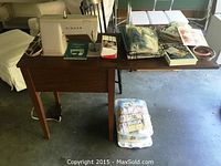 Wide view of the Singer sewing table with machine, sewing pedal underneath, instruction manual, buttonholer box, and various sewing supplies on top and below the table