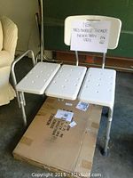 Med-Mobile shower chair viewed from front with white plastic seats and backrest above a cardboard box with product label.