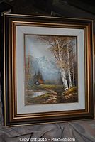 Framed fall landscape painting showing a scene with birch trees, a stream, a cabin, and mountains, with a decorative gold frame and white matting.
