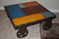 Full view of square table showing multicolored metal panel top, riveted steel frame, and caster wheels.