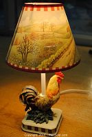 Table lamp showing the rooster base with the scenic countryside lampshade on top, lit to show warm glow