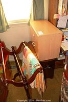 Wooden quilt rack holding a multicolored quilt next to light wood storage drawers and folding table.