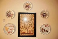 Wall arrangement showing all five decorative plates in a semi-circle around the framed antique cross stitch sampler. Each plate varies in design with historical, Asian, floral and pastoral themes visible.