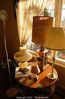 Group photo of six table lamps of various heights and one tall standing lamp near a window showing lamps' styles and materials.
