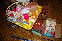 Overview of the children's floral table filled with various toys and craft items including a red and yellow lamp toy, plastic bags of craft material, boxes, and small toys.