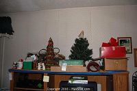 Wide view of a shelf showing a collection of Christmas holiday decor including two small trees, stacked boxes, ornament containers, and figurines.