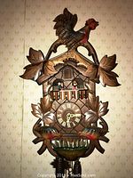 Front view of the cuckoo clock showing the carved rooster on top, leaf decorations, the clock face with Roman numerals, and two figurines in the window.