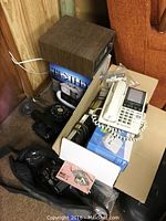 Photo of black rotary dial phone, beige rotary phone with buttons, and miscellaneous boxed accessories in corner of room.