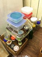 Stacked clear and colored lidded plastic containers with various glass jars in cardboard box.