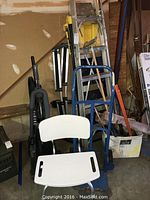 Photo showing a variety of items including a white shower chair with backrest, a blue hand truck, vacuums, and ladders stacked behind them.