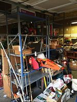 View of two large metal shelving units in a cluttered garage with various boxes, fishing rods, and miscellaneous items stored on them. Shows bent and rusty metal shelving surfaces and frame.
