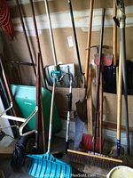 Wide view of garden tools stacked against wall showing many wooden handle tools, green wheelbarrow, blue shovel, broom, rakes, and spade.