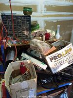 Photo of assorted hardware and other items piled on a table including a multi drawer small parts organizer, cardboard box of screws and nails, coil of wire, green rolls of tape, and a ProLine motor oil box.