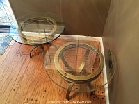 Pair of round end tables with clear circular glass tops and brushed metal bases, shown on wood floor near wall.