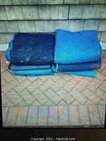 Photo showing 6 large folded moving blankets in different shades of blue, stacked against a couch on a carpeted floor.