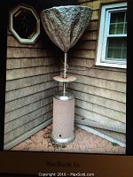 Photo of a tall patio heater with a propane tank base and two-tone tan and stainless steel finish, including a protective cover loosely fitted over the top reflector and upper unit.