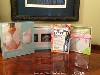 Photo showing all items together on a wooden surface under a framed picture on the wall: Belly casting kit box, sonogram frame with three slots, pregnancy book, feeding and diapering chart tied with a pink ribbon.