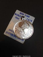 Front view of the sterling silver charm attached to original official New York World's Fair 1964-1965 souvenir packaging, showing embossed globe and text details.