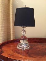 Glass bubble table lamp with black fabric shade shown from a side angle on a wooden surface against a light wall with curtain.