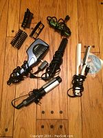 Multiple hair styling tools arranged on wooden floor: RED ceramic styler with attachments, black curling iron, and two silver flat irons.