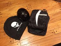 Photo showing black Fulmer motorcycle half helmet and folded black rain suit with white reflective panels next to a black dust bag with skull logo and 'FULMER' text on hardwood floor.