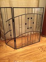 Full front and side view of the three-panel fire screen standing on a wooden floor with a beige textured wall background.