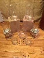 Photo showing set of 7 glass hurricane candle holders arranged on wooden floor, including 3 large and 4 medium holders with some original boxes visible.