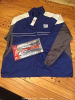 Full view of the Giants windbreaker jacket laid flat on a wooden floor with the Giants license plate frame placed on top, both new and unused.