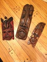 Photo showing all three hand carved African wooden masks on wooden floor; includes the Maasai warrior mask, elongated tribal head, and smaller mask.