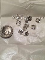 Photo of 15 loose white sapphires in assorted round and square cuts with a U.S. dime for size comparison on a white textured surface.