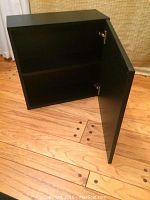 View of black square hanging cabinet with door open showing interior shelf and wood grain texture on door.