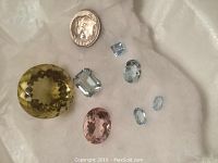 Photo showing seven loose faceted gemstones on white fabric with a dime for scale, including a large round yellow-green stone, a rectangular light blue stone, an oval peach-pink stone, a square pale blue stone, and three smaller light blue stones.